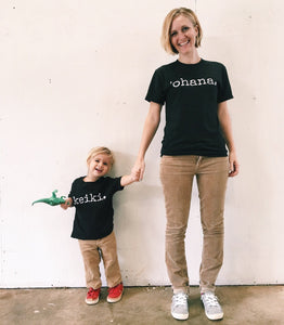 mother and son wearing matching tan pants and black tshirts with white writing and boy is carrying a dinosaur and wearing red shoes