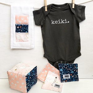 dark grey baby Ivy & Co. onesie with white lettering that says keiki hanging on a clothes line with pink burp cloth and baby soft block