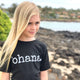 blonde girl standing on the rocks by the ocean wearing a black 'ohana tshirt