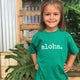 girl with hands on hips in front of monstera leaves wearing a green tshirt that says aloha