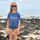blonde girl standing on the rocks by the ocean wearing sunglasses wearing a blue 'ohana tshirt