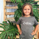 girl with hands on hips in front of monstera leaves wearing a grey tshirt that says aloha